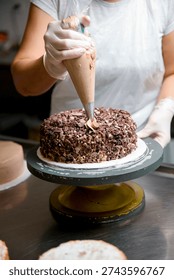 A baker is working in a kitchen. They use a piping bag to add icing to a chocolate cake. Other cake layers are placed nearby on a table.