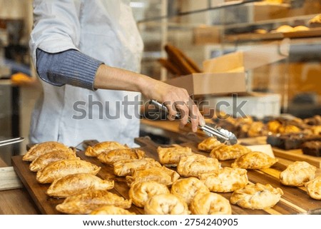 Baker's hand using tongs to select a freshly baked savory empanada from a wooden tray displayed in a glass bakery case, offering delicious food and service