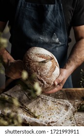 The Baker's hands hold a loaf of rye bread over a wooden table.