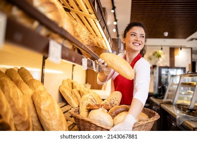 Bakery female worker in uniform selling loaf of bread to the customer in bakehouse. In background shelf full with fresh pastry products for sale.