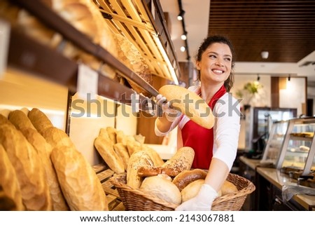Bakery female worker in uniform selling loaf of bread to the customer in bakehouse. In background shelf full with fresh pastry products for sale.