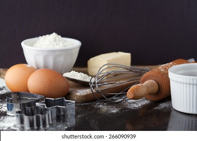 Baking cake ingredients with raw eggs, rolling pin, flour and cookie cutters on black background