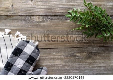 Baking concept flat lay, with black and white oven mitt, towel, and green leaves on a wood background