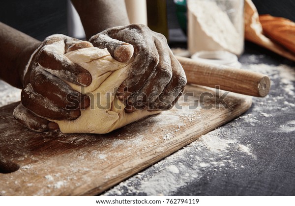 Baking concept. Hard working unrecognizable black male prepares pastry by himself, kneads dough on wooden counter with flour and rolling pin. Male African American cook bakes bread or delicious bun