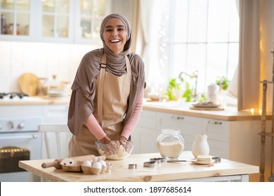 Baking Concept. Portrait Of Joyful Muslim Woman In Hijab Kneading Dough In Kitchen Interior, Cheerful Islamic Female In Hijab And Apron Having Fun While Preparing Homemade Pastry, Laughing At Camera