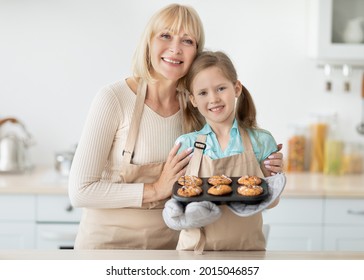 Baking Concept. Portrait of smiling grandmother embracing cute little girl who holding tray with freshly baked cookies homemade cakes. Smiling aged lady teaching kid how to cook, family wearing aprons - Powered by Shutterstock