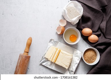 Baking Cooking Ingredients: A Bag Of Flour, Eggs, Egg With 
Yolk In A Bowl,  Brown Sugar, Butter Dark Linen Cloth, Rolling Pin, On Light Grey Concrete Background. Top View Copy Space.  Recipe Mockup