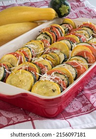 A Baking Dish Of Alternating Zucchini, Yellow Squash, And Tomato Circles, Topped With Parmesan Cheese And Garnished With Fresh Rosemary And Thyme Sits On A Red And White Table Covering