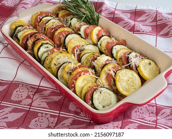 A Baking Dish Of Alternating Zucchini, Yellow Squash, And Tomato Circles, Topped With Parmesan Cheese And Garnished With Fresh Rosemary And Thyme Sits On A Red And White Table Covering