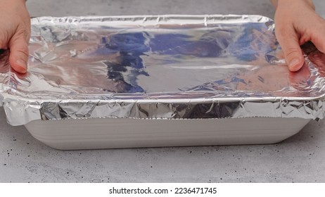 Baking dish covered with aluminum foil close up on the table, front view, woman hands. Spinach lasagna recipe. Before baking cover lasagna with aluminum foil. - Shutterstock ID 2236471745
