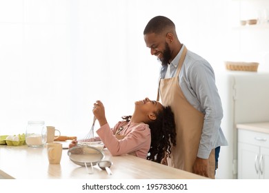 Baking With Father Is Fun. Happy Black Little Girl Mixing Flour And Dough In Bowl And Looking Up At Her Dad. Family Enjoying Cooking Homemade Food, Making Pastry In Kitchen Together, Copy Space