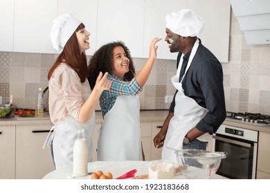 Baking Is Fine And Funny. Happy Curly Mixed Race Teen Girl Smutching Her Father's Face With Flour, While Having Fun With Her Parents In The Kitchen, Baking Together