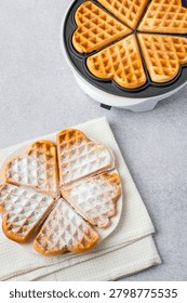 Baking heart-shaped Belgian waffles using an electric waffle iron and several waffles on a plate. 