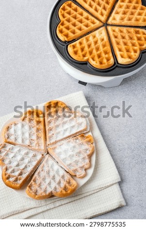 Baking heart-shaped Belgian waffles using an electric waffle iron and several waffles on a plate. 
