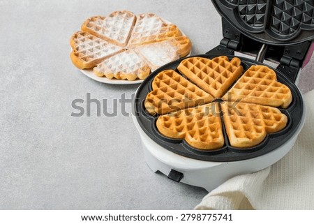 Baking heart-shaped Belgian waffles using an electric waffle iron and several waffles on a plate. Horizontal shot from above.