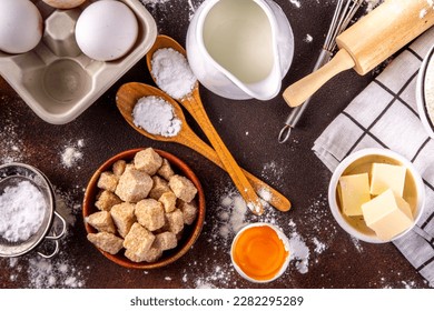 Baking ingredients background. Cooking ingredients (flour, eggs, milk, brown sugar, butter) with utensils on dark brown background top view copy space