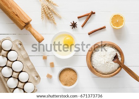 Baking ingredients: flour, eggs, brown sugar, spices, lemon, rolling pin and golden wheat ears on white wooden table. Top view