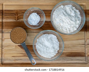 Baking Ingredients In Glass Bowls And A Metal Measuring Cup Overhead View On A Wooden Board. Brown Sugar, Baking Soda, Flour And Sugar. 