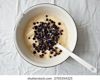 Baking process, making a berry cake: frozen berries added to a mixing bowl with vanilla cake batter. Bowl is white enamelled metallic with black rim, white plastic mixing spatula. White tablecloth.