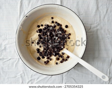 Baking process, making a berry cake: frozen berries added to a mixing bowl with vanilla cake batter. Bowl is white enamelled metallic with black rim, white plastic mixing spatula. White tablecloth.