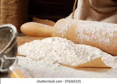 Baking still-life with flour heaped onto parchment paper, vintage wooden rolling pin, vintage sieve, and canvas flour sack with rope in background.  Closeup with shallow dof.  Selective focus on flour