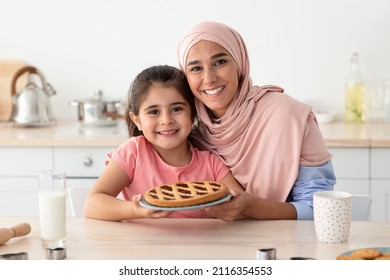 Baking Together. Portrait Of Happy Arabic Mother And Little Daughter Holding Homemade Pie And Smiling At Camera While Sitting At Table In Kitchen, Cheerful Islamic Family Enjoying Cooking At Home