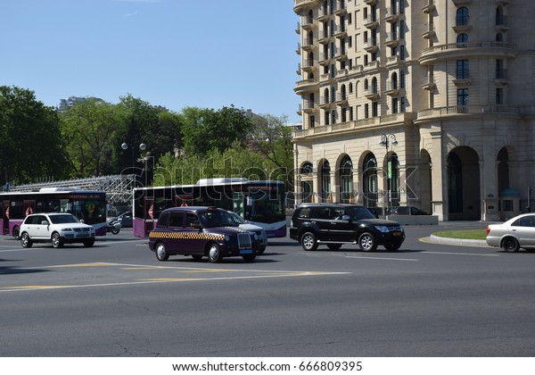 Baku, Azerbaijan -\
May 07, 2017. Two buses in white and maroon-violet livery and a\
taxi cab pass a square