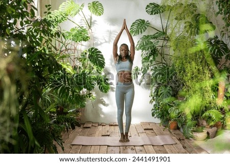 Balanced african american woman standing on fitness mat with closed eyes, hand together above head focusing on breath doing yoga surround by urban jungle interior. Relaxed black female calm meditation