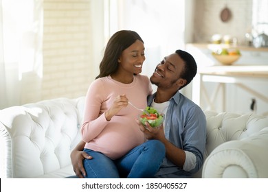 Balanced Diet In Pregnancy Concept. Handsome Black Man Giving His Lovely Expectant Wife Yummy Vegetable Salad Indoors. Positive Black Couple Eating Healthy While Waiting For Baby