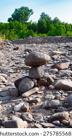 A balanced rock cairn or stone stack sitting in the middle of a dry, rocky riverbed. The background features a lush line of green trees under a soft blue sky. The image conveys a sense of peace, balance, Zen, and human connection to the natural landscape.