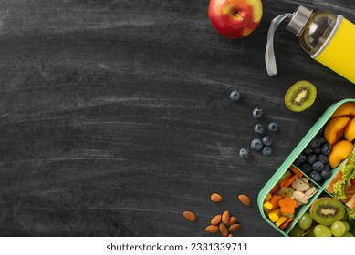 Balanced snack idea for school. Top-down view showcasing plastic lunchbox packed with fresh fruits, sandwich, berries, nuts, water bottle, placed on blackboard backdrop, space for text or ad content