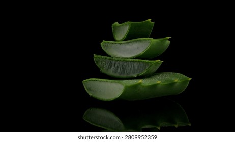 A balanced stack of sliced aloe vera leaves reveals the translucent, succulent inner gel. Dramatic side lighting highlights the vibrant green skin and moist texture against a reflective black background.