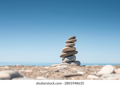 Balanced stack of smooth pebbles on a quiet beach with blue sky and sea horizon. Minimalist coastal scene symbolizing balance, harmony, mindfulness, stability and inner peace. Relaxation, mindfulness, self-care, and personal development concepts. Copy space