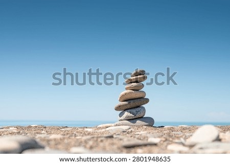 Balanced stack of smooth pebbles on a quiet beach with blue sky and sea horizon. Minimalist coastal scene symbolizing balance, harmony, mindfulness, stability and inner peace. Relaxation, mindfulness, self-care, and personal development concepts. Copy space