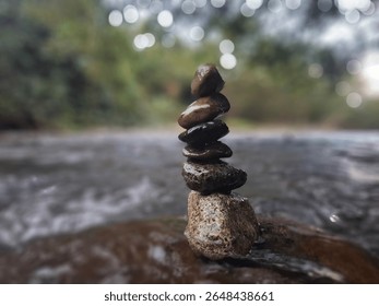 A balanced stack of smooth, wet river stones stands tall amidst the flowing water, with a blurred natural background and bokeh lights