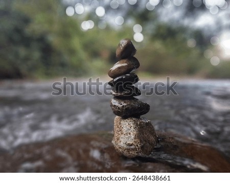 A balanced stack of smooth, wet river stones stands tall amidst the flowing water, with a blurred natural background and bokeh lights