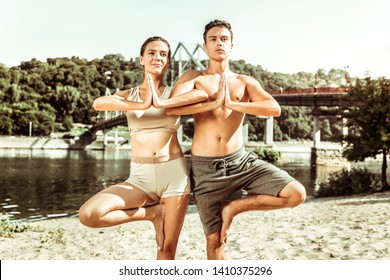 Balancing Asana. Closeup Of A Nice Smiling Girl Standing On The Beach While Practicing A Tree Pose With Her Young Partner