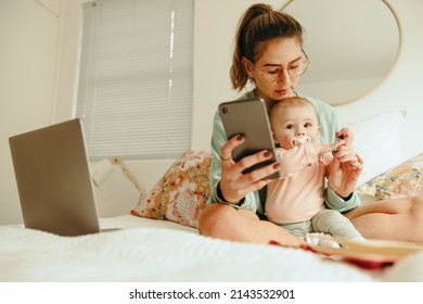 Balancing Motherhood And Work. New Mom Taking A Pen From Her Baby While Working On A Bed. Multitasking Single Mother Trying To Read A Text Message While Working From Home.