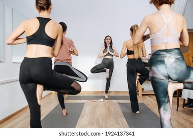 Balancing on one foot young women practicing yoga in a group in front of an instructor. They are standing on one foot, second foot against thigh, joining hands. View from behind backs.
