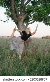 Balancing On One Foot Young Blond Woman Exercising Under A Tree On Grass. She Is Wearing Skirt Pants And Black Shirt. Low Angle.