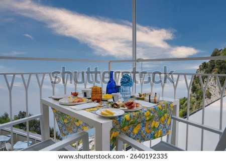 A balcony with a lemon patterned tablecloth, breakfast items, and modern furniture overlooks the Tyrrhenian Sea, with greenery and a clear sky.