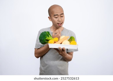 A bald Asian man who looks scared and hesitant when looking at a tray filled with a variety of fresh vegetables and fruits, a campaign to consume healthy and nutritious food, the concept of a healthy.