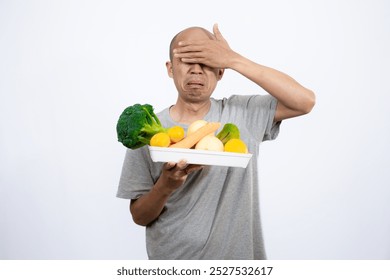 A bald Asian man who looks scared and hesitant when looking at a tray filled with a variety of fresh vegetables and fruits, a campaign to consume healthy and nutritious food, the concept of a healthy.