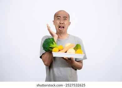 A bald Asian man who looks scared and hesitant when looking at a tray filled with a variety of fresh vegetables and fruits, a campaign to consume healthy and nutritious food, the concept of a healthy.