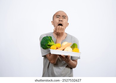 A bald Asian man who looks scared and hesitant when looking at a tray filled with a variety of fresh vegetables and fruits, a campaign to consume healthy and nutritious food, the concept of a healthy.