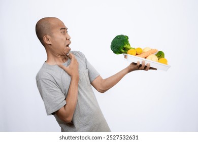 A bald Asian man who looks scared and hesitant when looking at a tray filled with a variety of fresh vegetables and fruits, a campaign to consume healthy and nutritious food, the concept of a healthy.