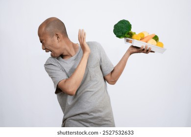 A bald Asian man who looks scared and hesitant when looking at a tray filled with a variety of fresh vegetables and fruits, a campaign to consume healthy and nutritious food, the concept of a healthy.