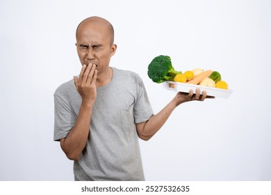 A bald Asian man who looks scared and hesitant when looking at a tray filled with a variety of fresh vegetables and fruits, a campaign to consume healthy and nutritious food, the concept of a healthy.