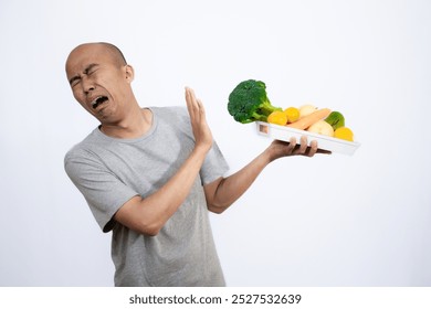 A bald Asian man who looks scared and hesitant when looking at a tray filled with a variety of fresh vegetables and fruits, a campaign to consume healthy and nutritious food, the concept of a healthy.