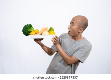 A bald Asian man who looks scared and hesitant when looking at a tray filled with a variety of fresh vegetables and fruits, a campaign to consume healthy and nutritious food, the concept of a healthy.